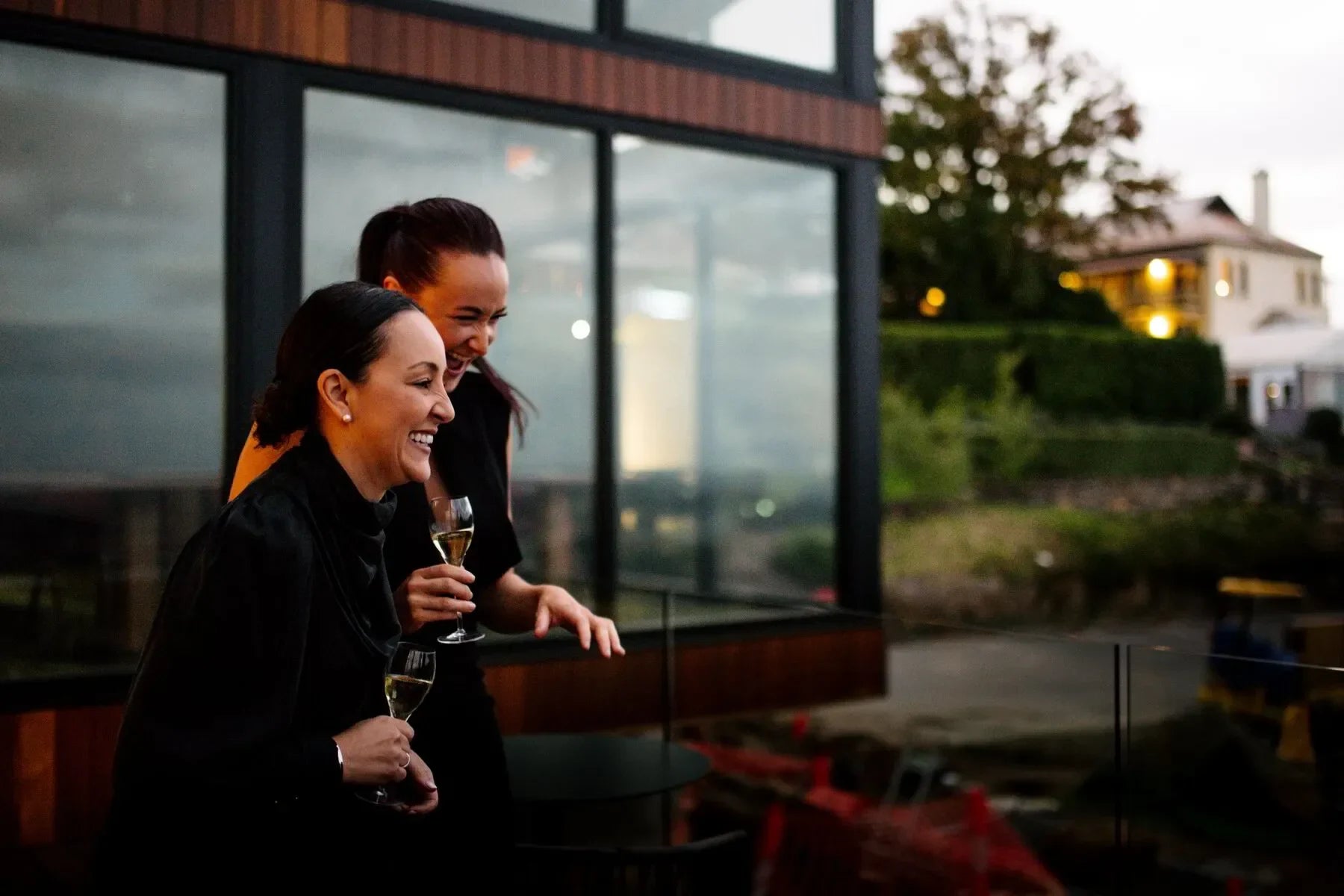 Two women laughing outdoors with wine glasses, modern building and greenery in background
