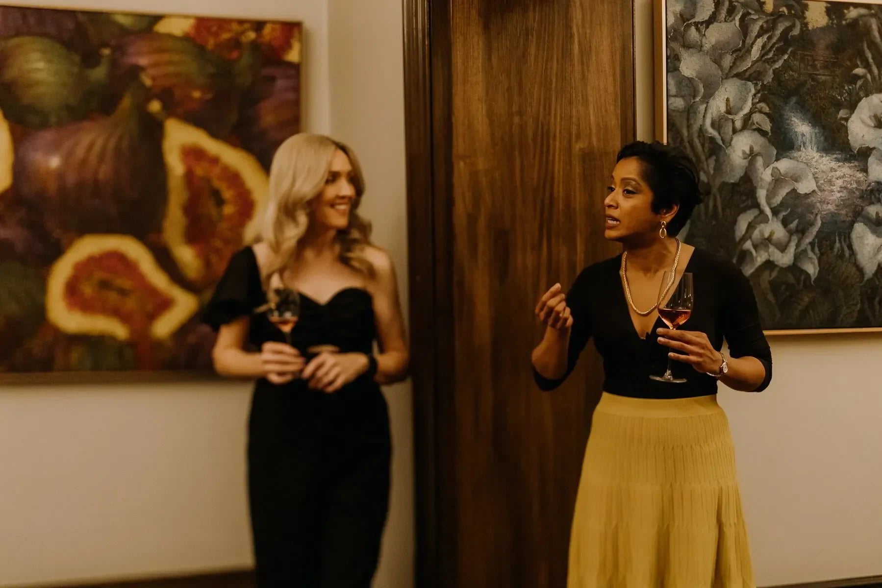 Two women holding wine glasses talking at an art gallery event with colorful paintings