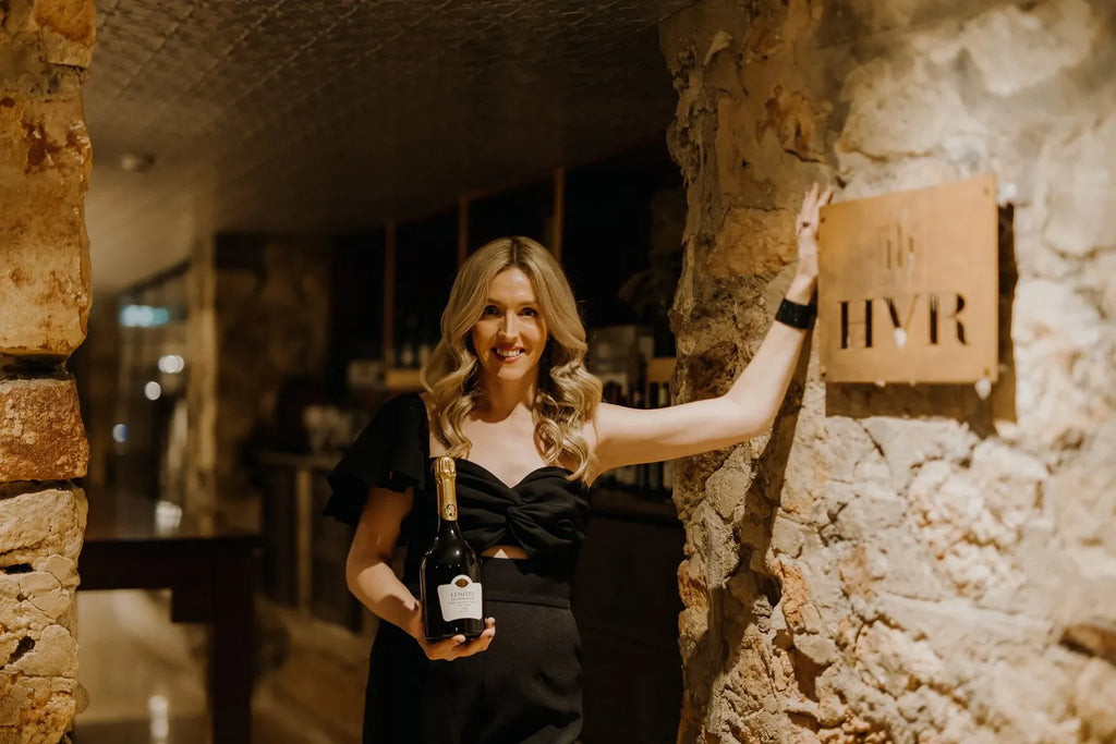 Woman in black dress holding champagne bottle in rustic wine cellar, HVR sign on stone wall