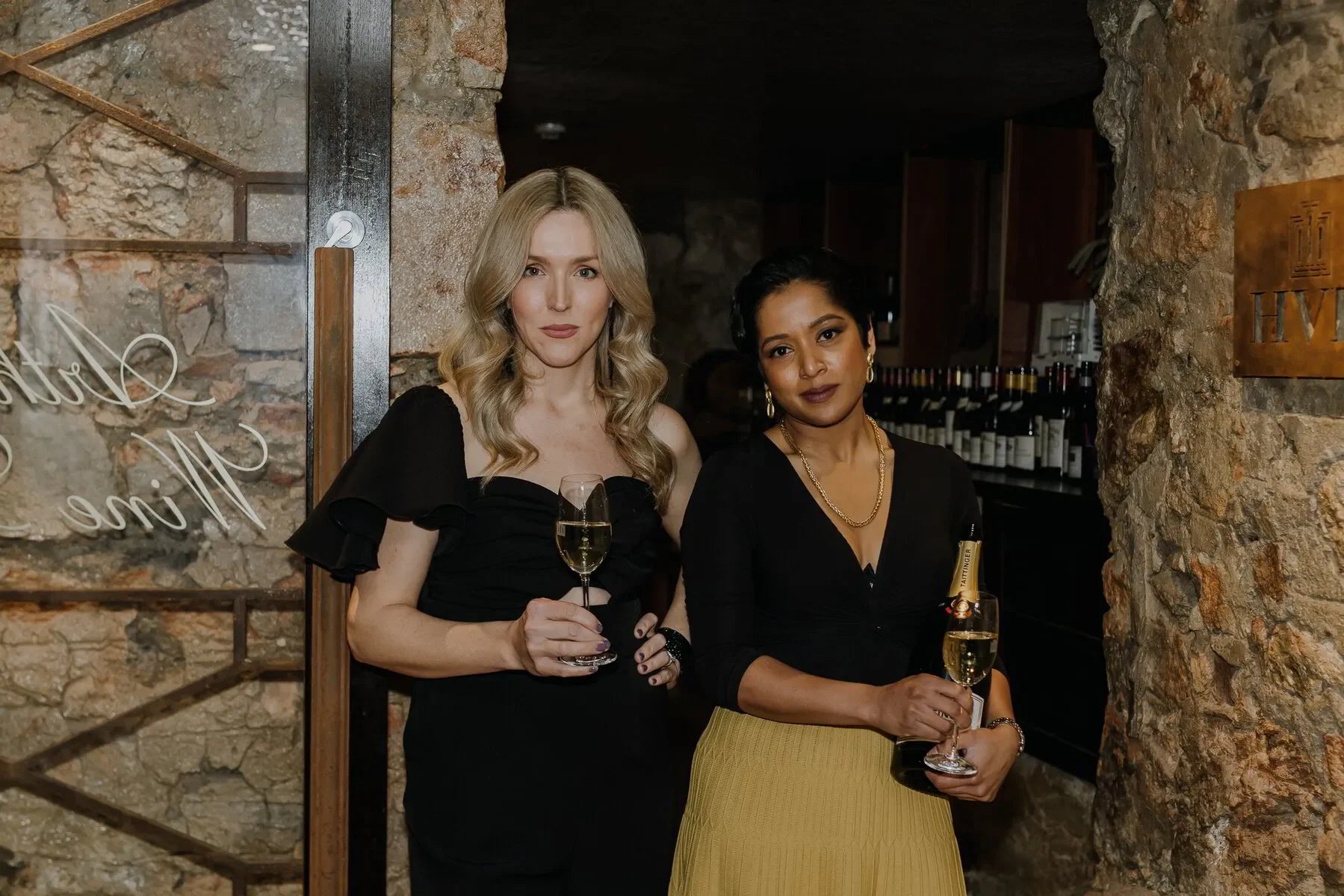 Two women holding champagne glasses at a wine cellar dinner event with stone walls.