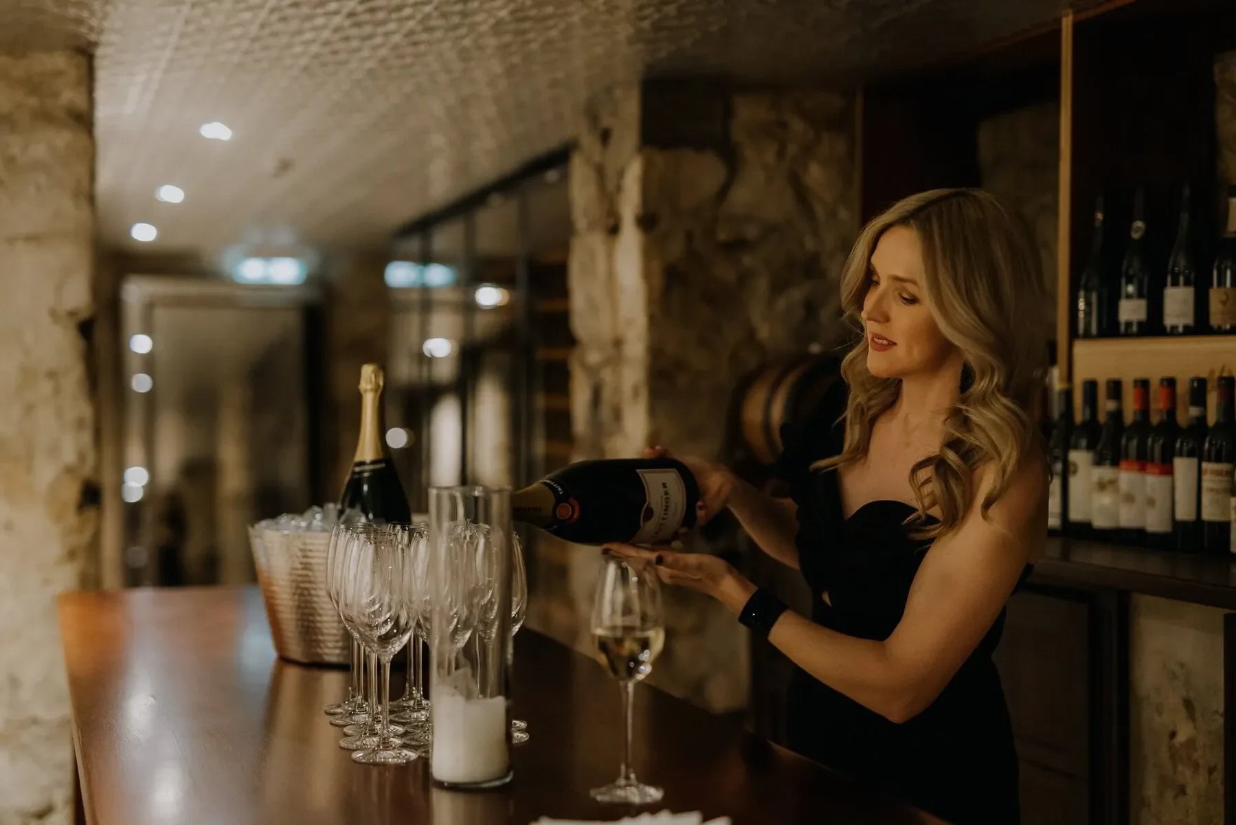 Woman pouring Taittinger champagne in wine cellar with glasses and wine bottles on counter