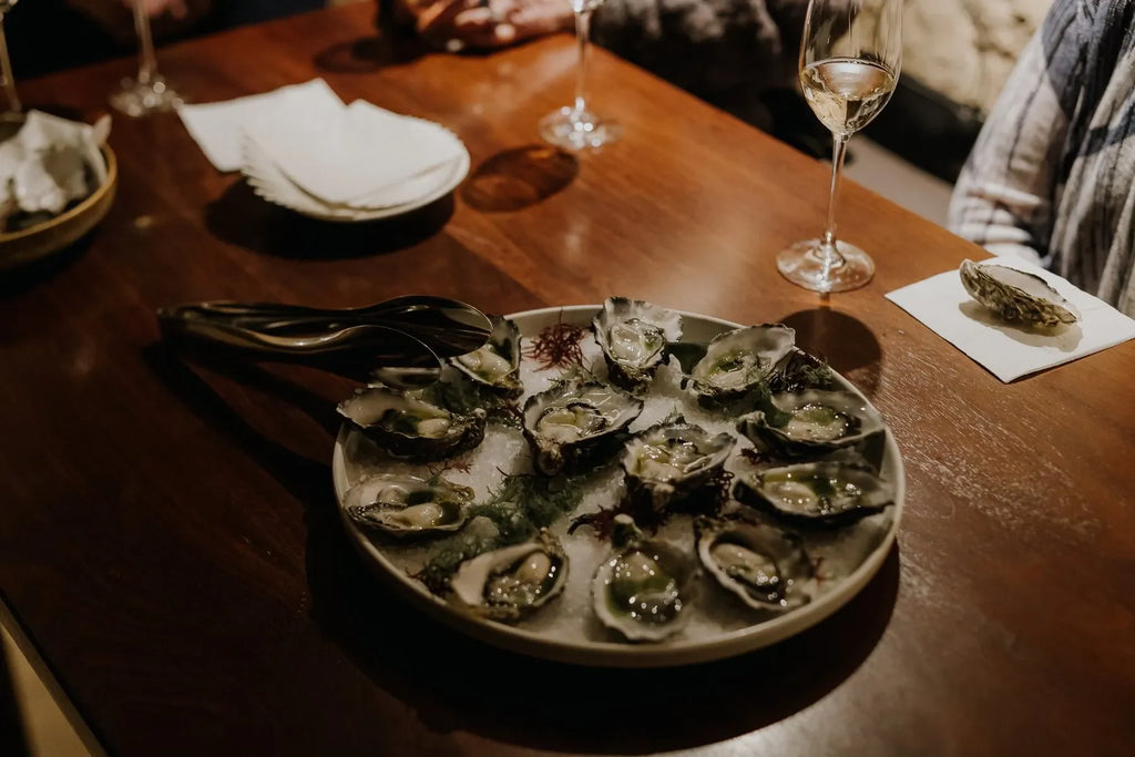 Plate of fresh oysters on ice with tongs, wine glasses, and napkins on wooden dining table