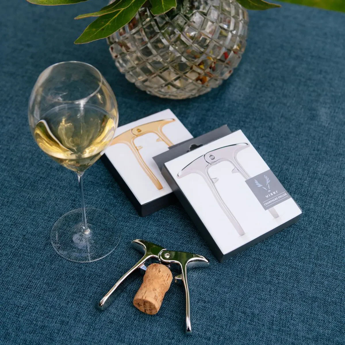 Viski champagne cork puller with packaging, wine glass and cork on blue tablecloth